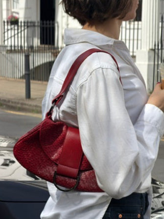 A person wearing a white button-up shirt and dark jeans carries a textured red shoulder bag outdoors on a street with buildings in the background.