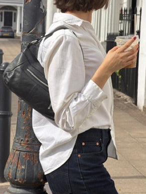A woman wearing a white long-sleeve button-up shirt and dark blue jeans stands on a city sidewalk next to a black metal pole, carrying a black shoulder bag and holding an iced coffee.