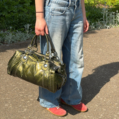 Person holding a green handbag outdoors on a paved path with greenery in the background