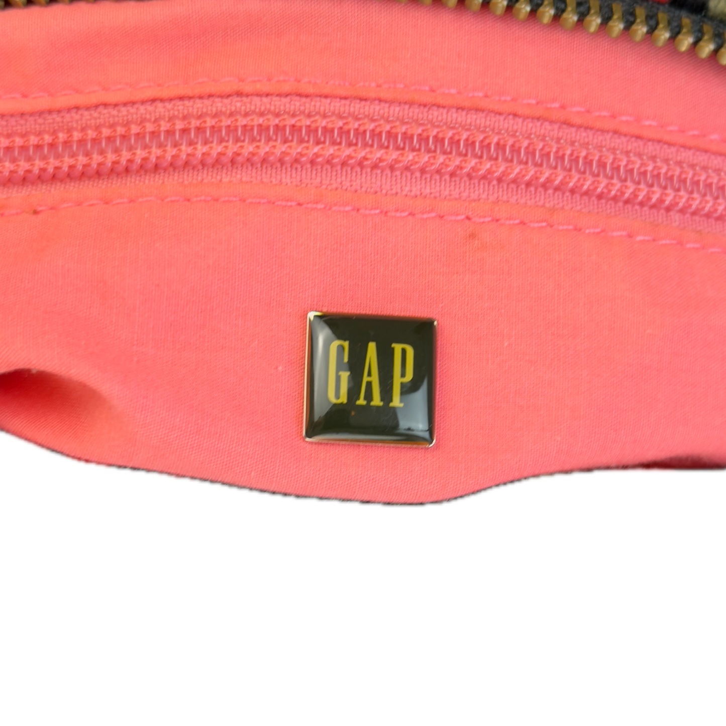 A close-up of a bag's bright pink interior with a gold-tone zipper, featuring a small square black and gold GAP logo patch on the front.