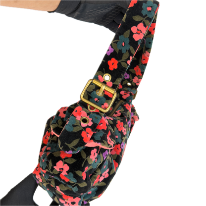 A close-up of a floral shoulder bag with a black velvet-like base and a dense print of pink, red, purple, and green flowers. The bag features a prominent gold-tone buckle and strap hardware, shown against a plain white background.