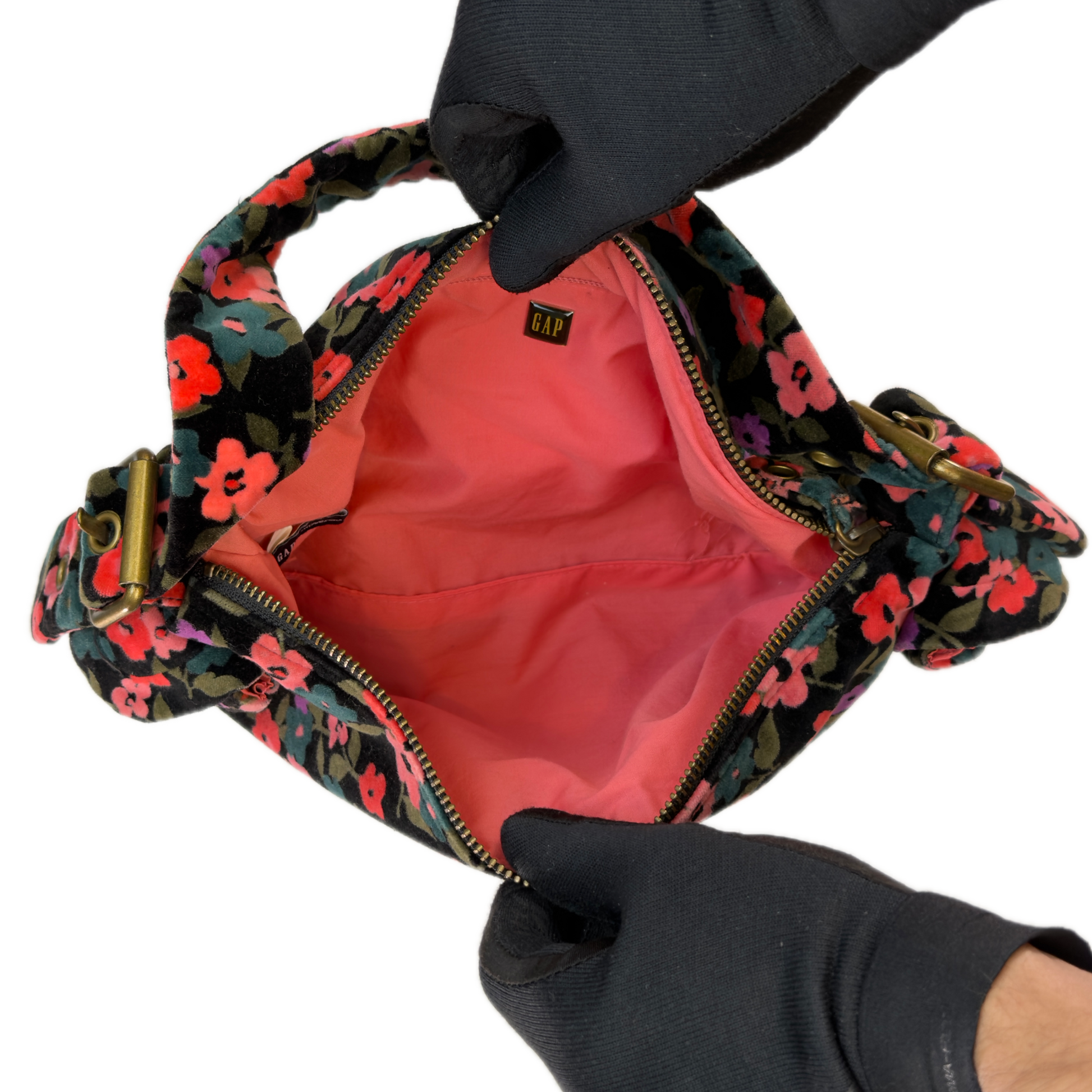 A close-up of a floral shoulder bag being held open by two gloved hands. The bag has a black base with a vibrant floral pattern in pink, red, and blue, and a bright coral-pink interior lining.