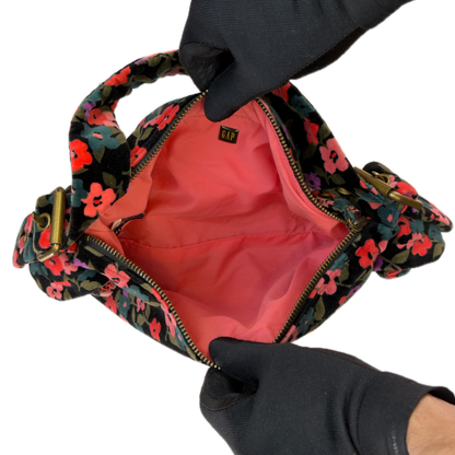 A close-up of a floral shoulder bag being held open by two gloved hands. The bag has a black base with a vibrant floral pattern in pink, red, and blue, and a bright coral-pink interior lining.