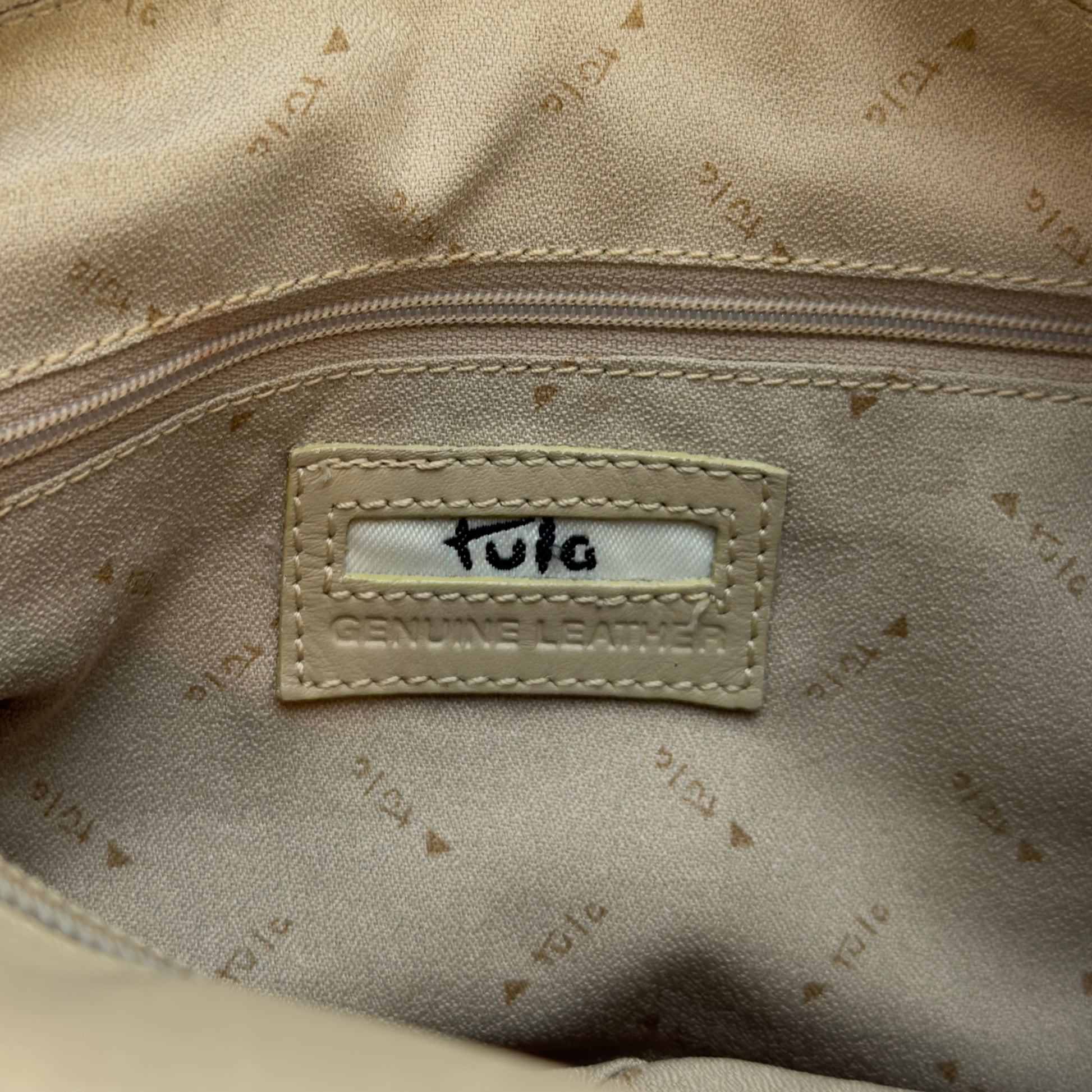 Close-up of the interior of a beige bag showing a stitched label that reads 'Tula' and 'Genuine Leather' on a patterned beige fabric.